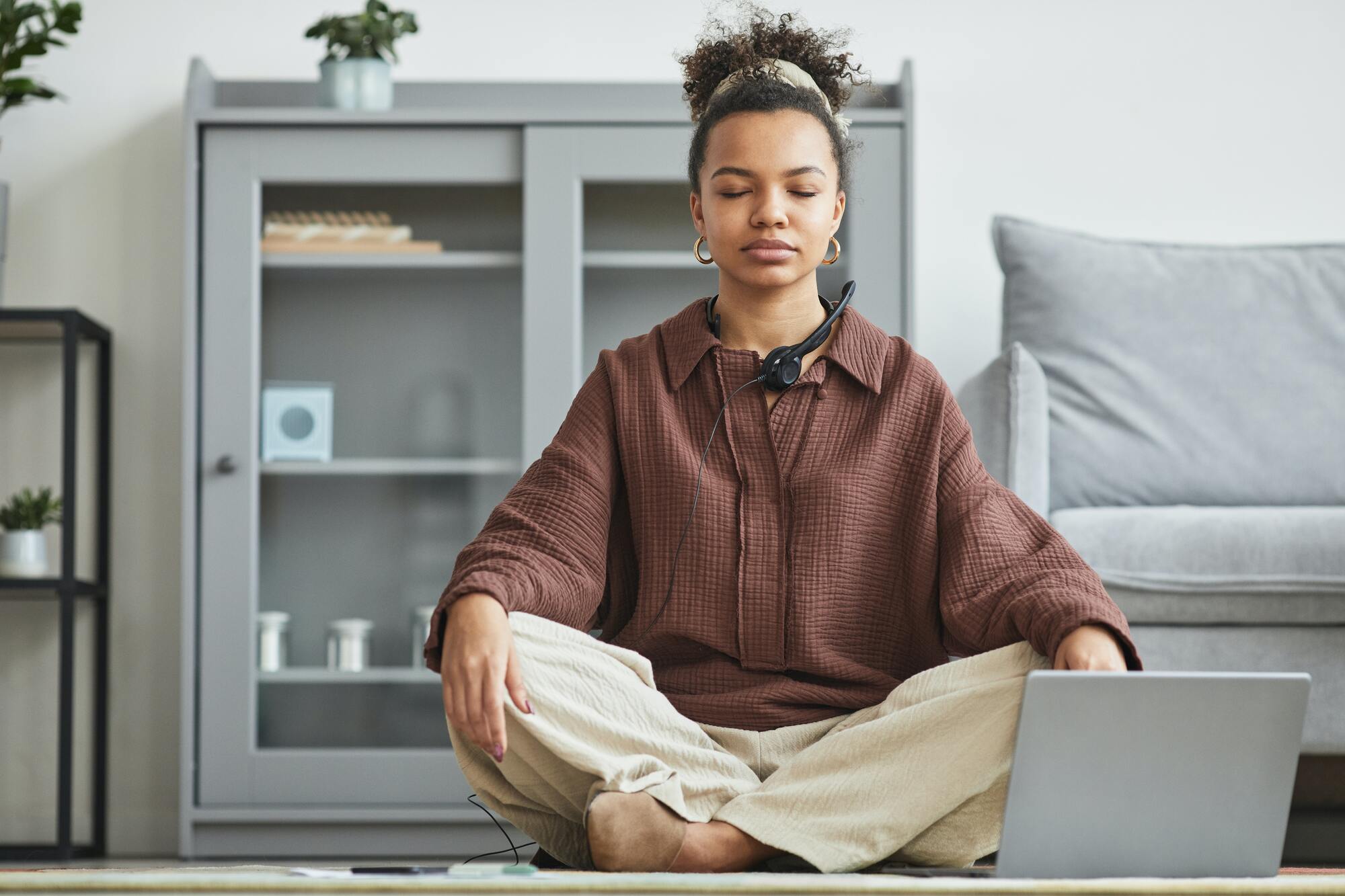 Woman sitting on her living room floor in front of her laptop meditating with her eyes closed.