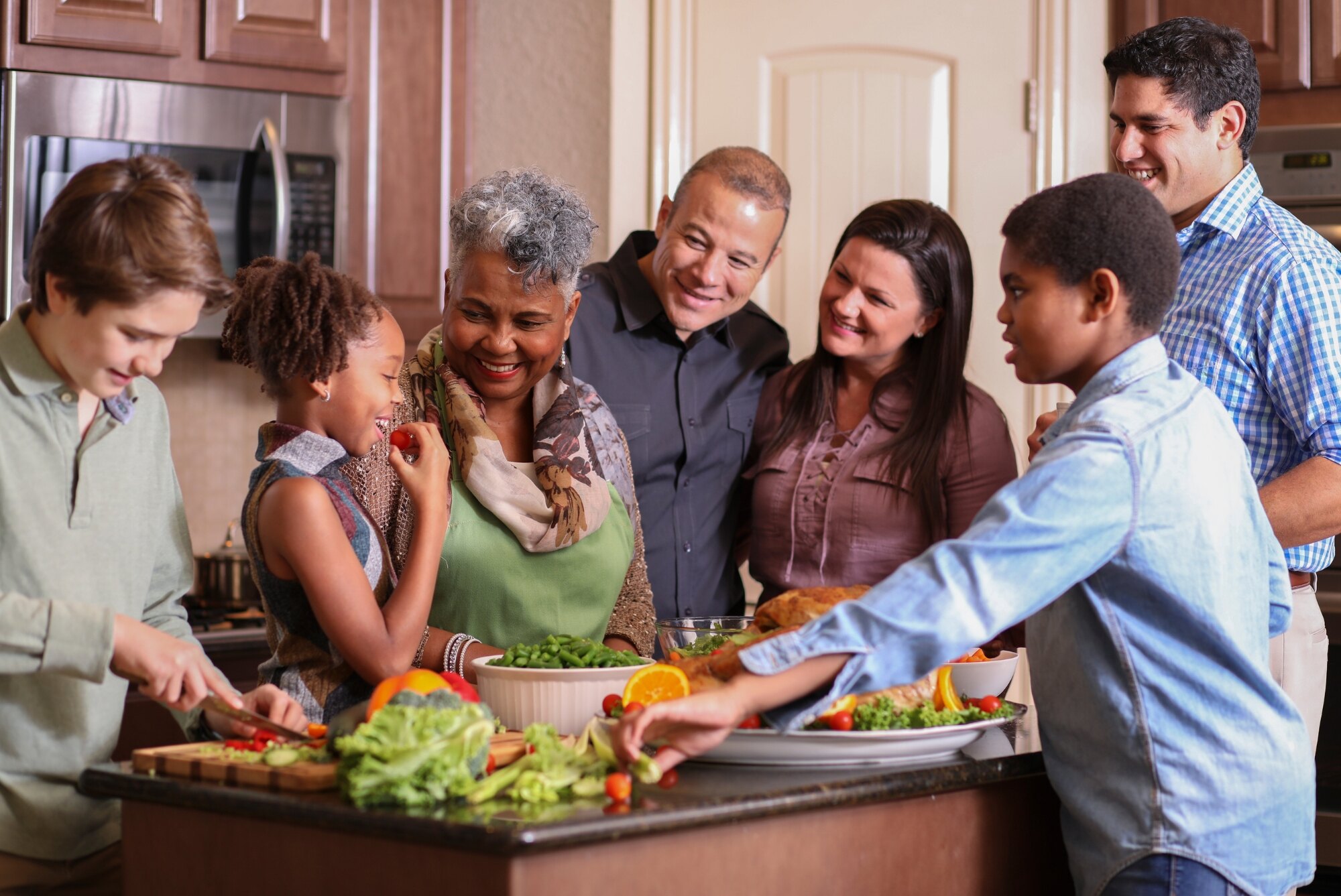 Diverse Family In Kitchen smiling as a young child bites into a fruit.